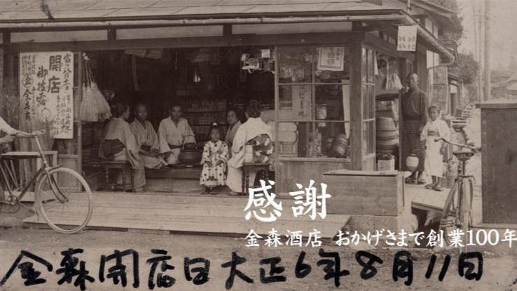 白黒の古い写真で、木造の店先に着物姿の人々が座り、傍らに自転車が停まり、天板や看板が掛かった板壁の店内がのぞく